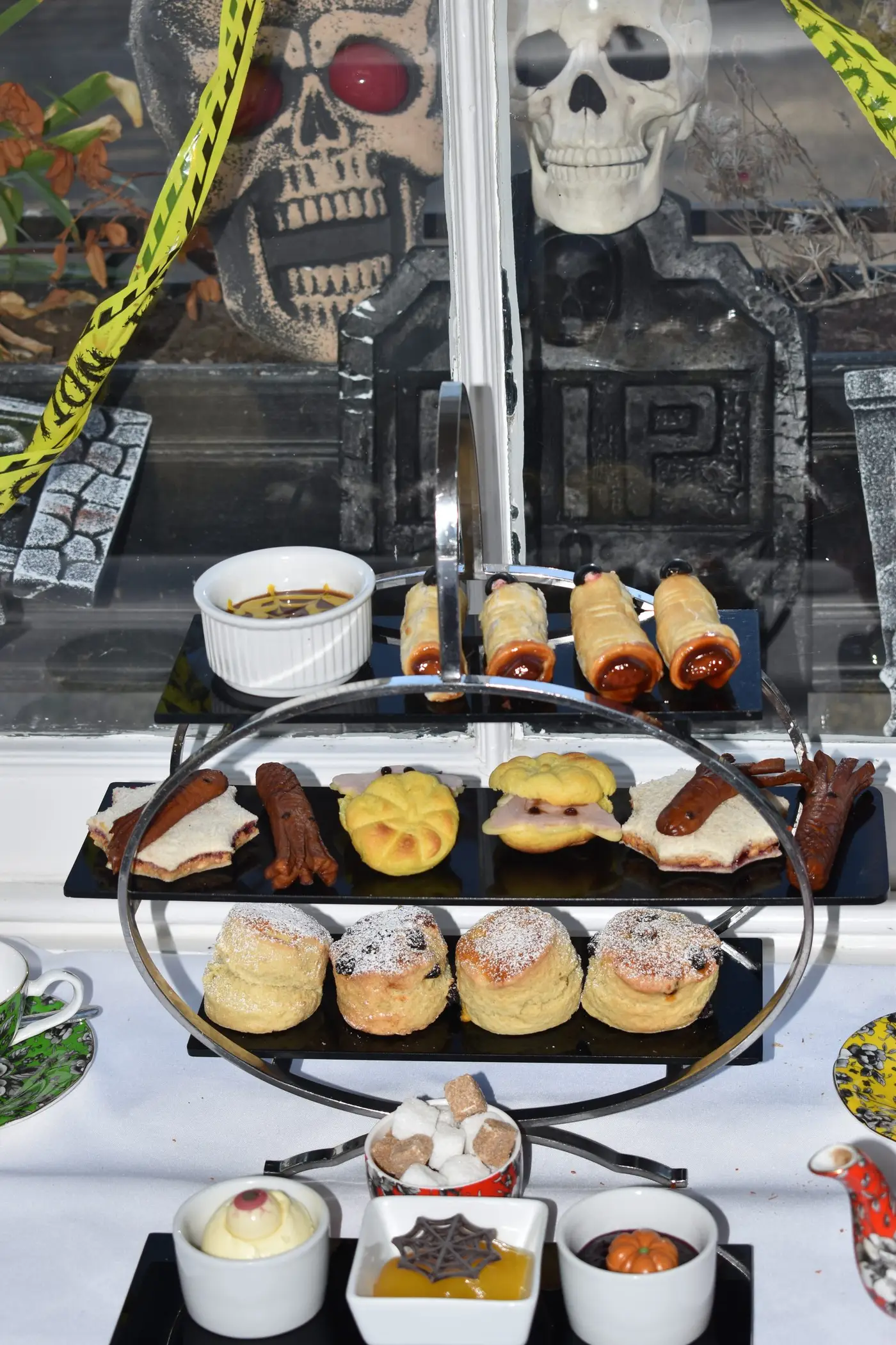Kids’ Halloween afternoon tea stand with finger sandwiches, pastry mummies and scones in front of a skull and gravestone display